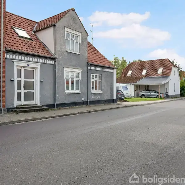 Gråt murstenshus med rødt tag står langs en stille gade. Andre huse ses i baggrunden under en blå, let skyet himmel. Parkerede biler er til stede i baggrunden.