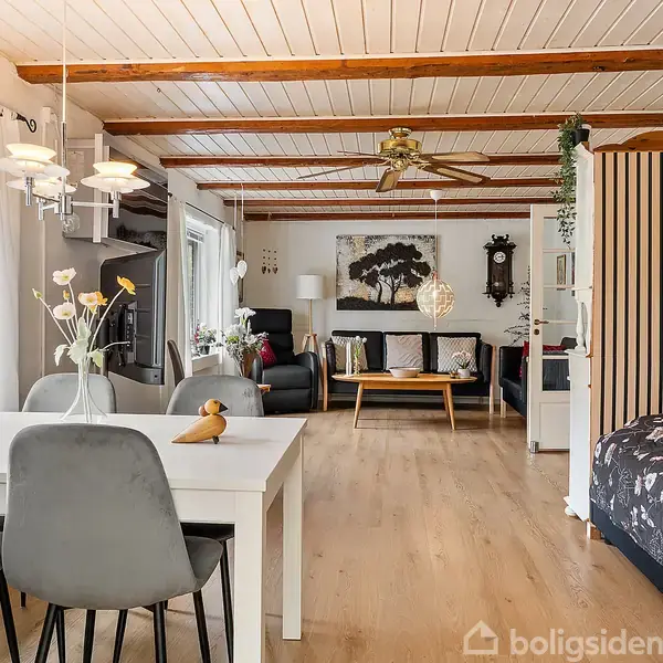 Stue i træhus har spisebord med fire stole, sofa, vægkunst og loftsventilator. Lysindfald fra store vinduer, blomsterdekorationer på bordet, trægulv og loftsbjælker.