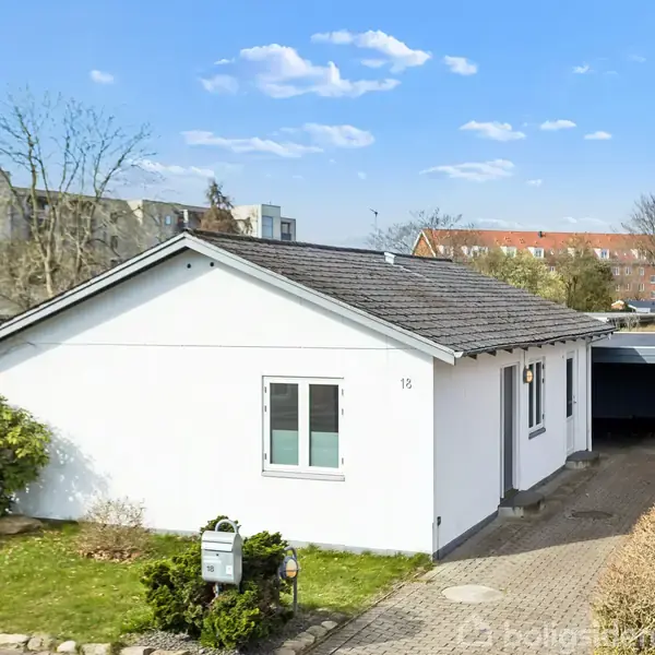 Hvidt enfamilieshus står stille i en have med en flisebelagt indkørsel. Husnummer 12 er synligt. Nabobygninger og træer ses i baggrunden under en blå himmel med skyer.