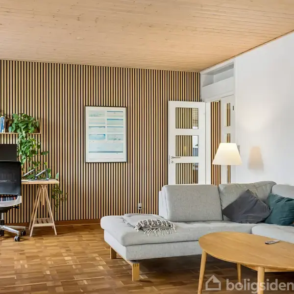 En stue med en grå hjørnesofa, sofabord og gulvlampe. Skrivebord med stol står op ad en væg med træpaneler. Rummet har et trægulv og dekoreret med grønne planter.