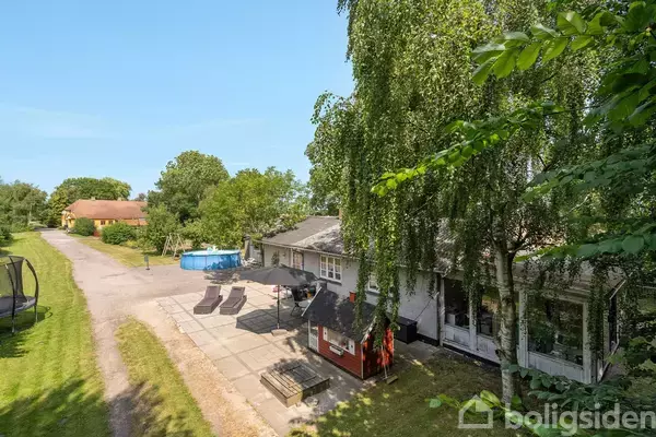 Hus omgivet af grønne træer og buske, med en gårdhave med liggestole og en børnepool på grusvej. Skyggefuldt område under store træer.