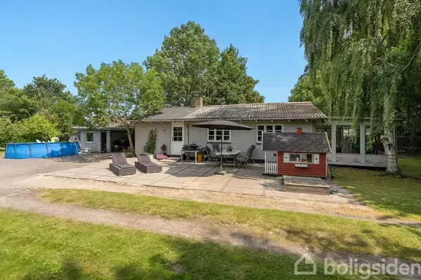 Et hus med fladt tag står omgivet af grønne træer og have, med en terrasse fyldt med havemøbler og et lille legehus foran. En blå swimmingpool ses til venstre.