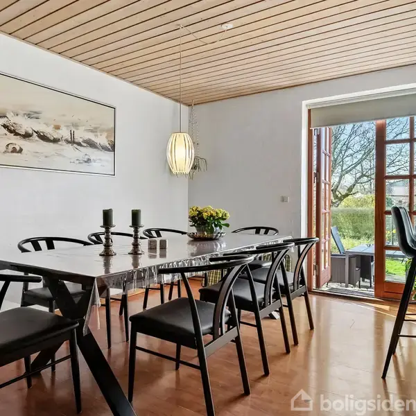 Et spisebord med sorte stole står i en spisestue med hvide vægge. En lampe hænger over bordet, og en glasdør fører ud til en have. En stor maleri hænger på væggen.