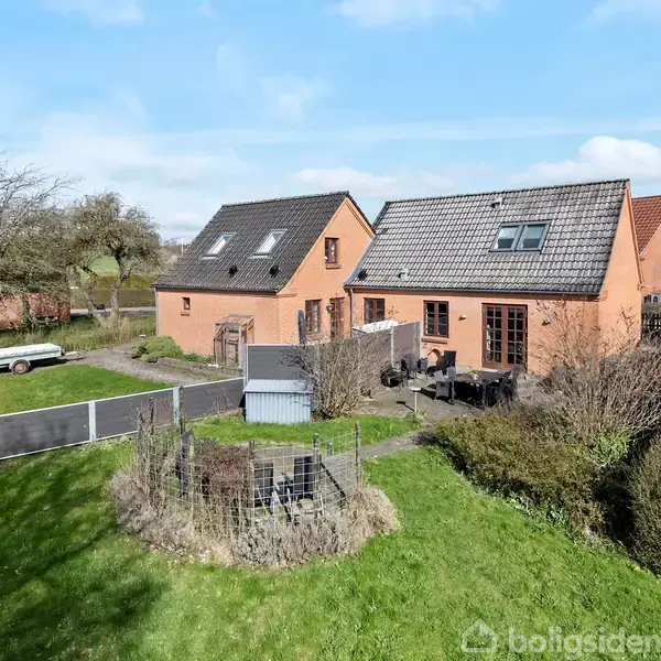 En rød murstensvilla med gråt tegltag og flere tagvinduer står i en velholdt have med grøn græsplæne og buske. En terrasse med havemøbler er placeret i nærheden.
