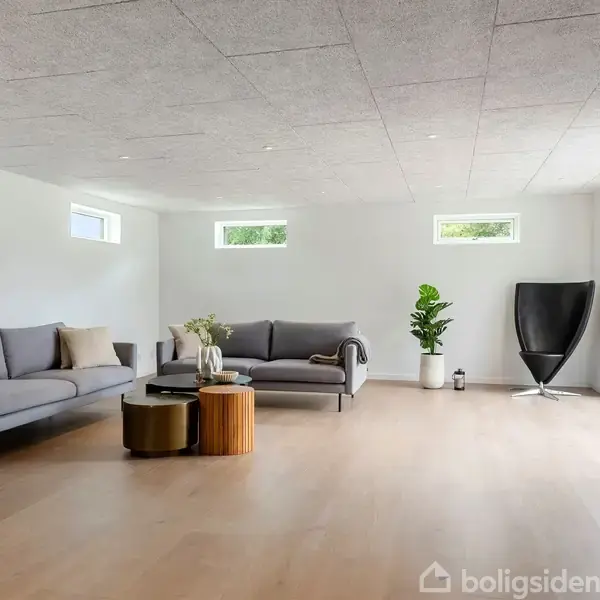 En rummelig stue med lysegrå sofaer står opstillet. Et sofabord har dekorationer. Store vinduer giver naturligt lys, og potteplanter samt en moderne stol supplerer det minimalistiske interiør.