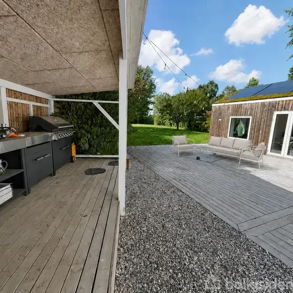 Udendørs køkkenområde til venstre med grill på træveranda; terrasse med sofa og stole til højre; græsklædt have og træer i baggrunden; klar himmel.