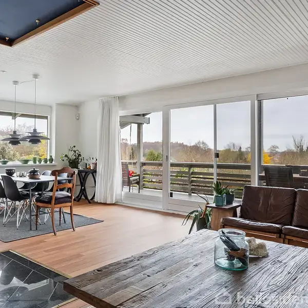 Stue med spisebord til seks personer ved et vindue, lædersofa og sofabord i forgrunden, udsigt til terrasse og have gennem store glasdøre.