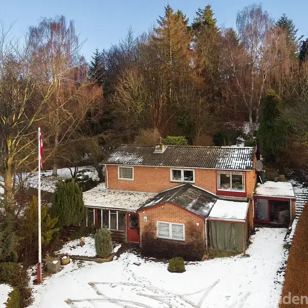 To-etagers murstenshus står stille i et snedækket haveområde med høje, nøgne træer omkring. En flagstang står i haven, hvilket skaber en vinterlig atmosfære.