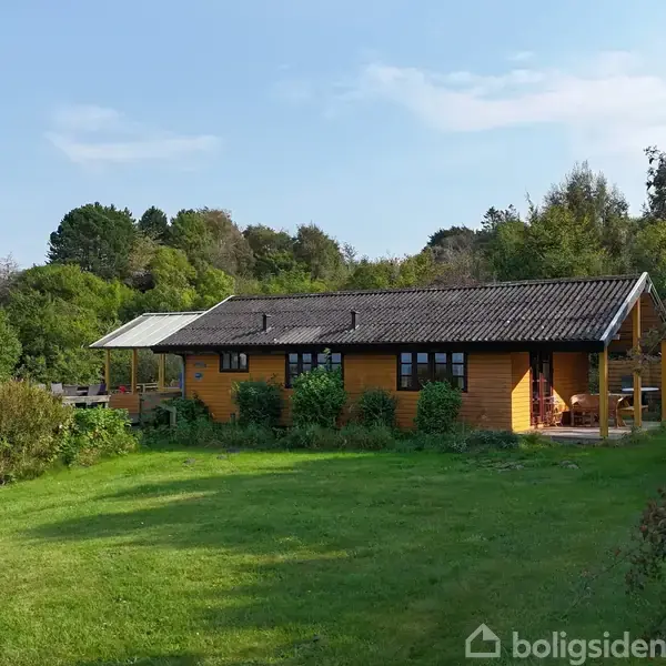 Et gul træhus står stille omgivet af en grøn have. Rundt om huset er der tætte buske og træer i en landskabelig og fredelig natur.