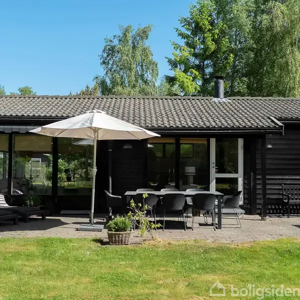 Et sort træhus står stille på en flisebelagt terrasse med havemøbler og parasoller. Grønne træer og buske omkranser huset under en klar blå himmel.