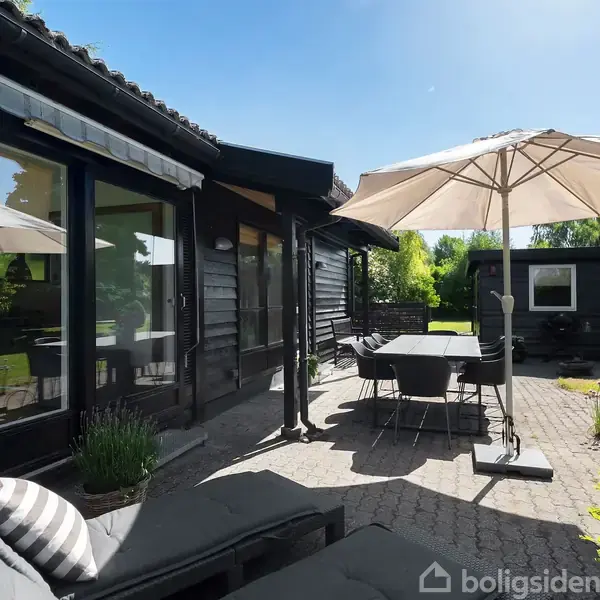 Udendørs terrasse indeholder en parasol, stole og bord i et stenbelagt område. Mørkt træhus tæt hertil. Sollys oplyser haven med grønne træer og buske.