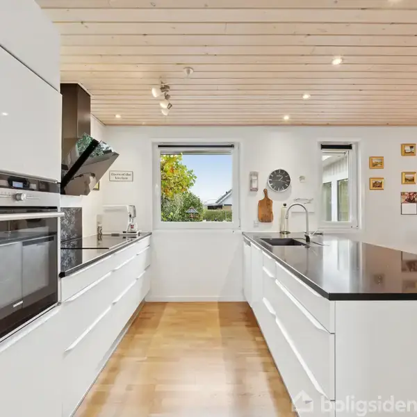 Et moderne køkken med hvide skabe og sort bordplade. Køkkenmaskiner er indbygget i væggen. To vinduer slipper udendørsbelysning ind. Trægulve og loftslamper skaber en lys atmosfære.