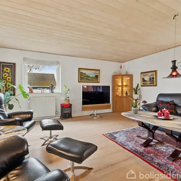 Stue med sorte lædermøbler på trægulv. Et sofabord står på et rødt tæppe. Vinduer og kunst på væggene tilføjer lys og farve. Nogen tekst er ikke synlig.