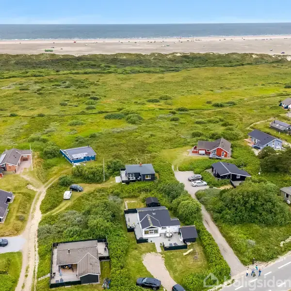 Sommerhuse ligger spredt blandt grønne buske nær en bred sandstrand i horisonten; en stille, landlig kystscene fanger det danske kystområdes charme.
