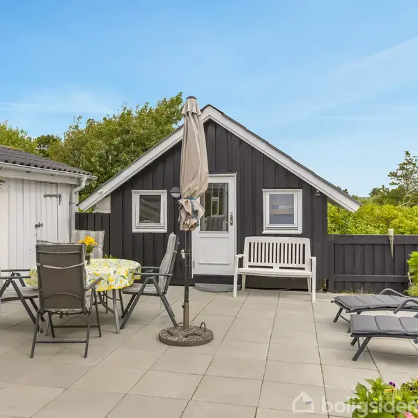 Et gårdhaveområde med fliser indeholder et bord med stole og en parasol. I baggrunden ses et sortmalet hus med en hvid dør og bænk. Træer omgiver området.