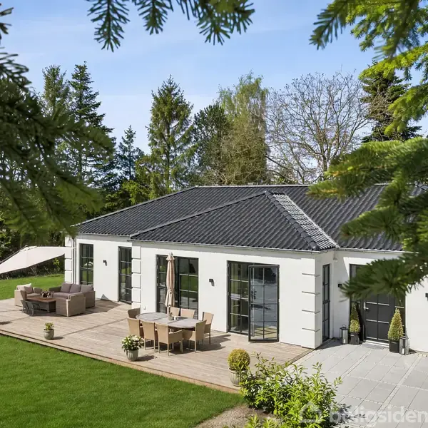Et moderne enfamiliehus med en terrasse. Der står havemøbler og en parasol på terrassen. Omgivet af en grøn have med træer i baggrunden.