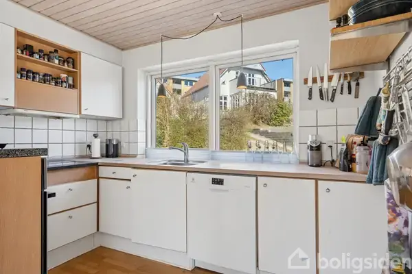 Et køkken har hvide skabe, en vask, knive på væggen, og krydderier på en hylde. Udsigten gennem vinduet viser en bakke med flere huse.