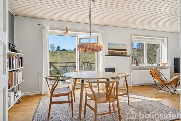 Et rundt spisebord med træstole står på et tæppe i et lyst rum. Store vinduer og en glasdør åbner mod en terrasse og græsareal udenfor. En hvid reol fyldt med bøger står i hjørnet.