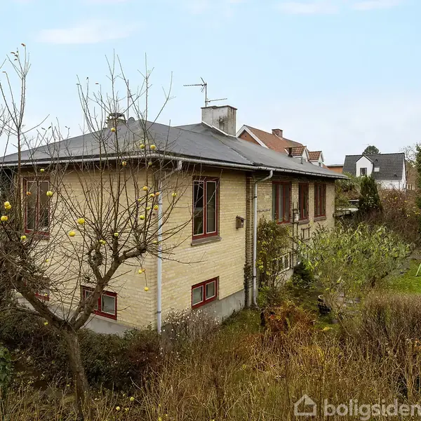 Et gulstenshus med røde vinduesrammer står i en have med frugttræer og en græsplæne. Huset ligger i et boligområde med flere omkringliggende huse.