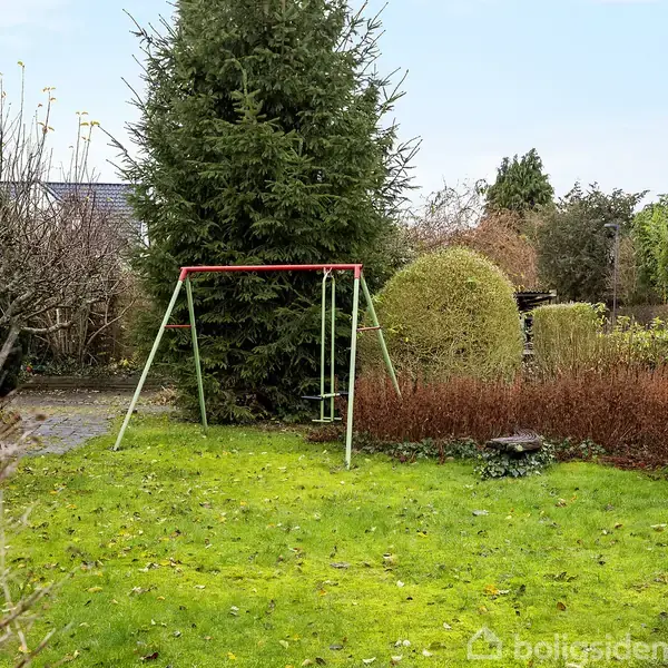 En gyngestativ står uden bevægelse på en grøn græsplæne omgivet af tæt beplantning og buske i en have.