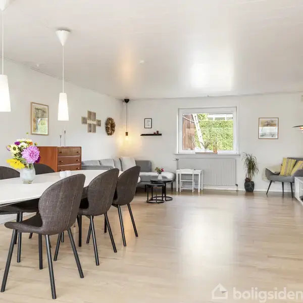En moderne spisestue med et langt bord og seks stole, blomsterdekoration i midten, lysende pendellamper ovenover og en sofa i baggrunden. Et vindue åbner mod en grøn have.