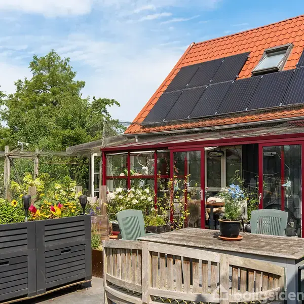Bord og stole står på en terrasse med blomster og planter omkring. Huset har solpaneler på det røde tag. Træer og en blå himmel danner baggrund.