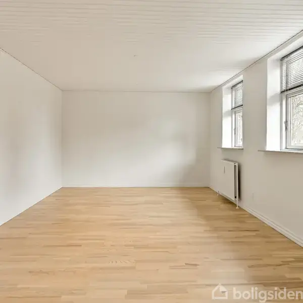 Tomt rum med trægulv og hvide vægge; to vinduer med radiator under i højre side. En lukket dør er synlig til venstre. Lys strømmer ind fra vinduerne.