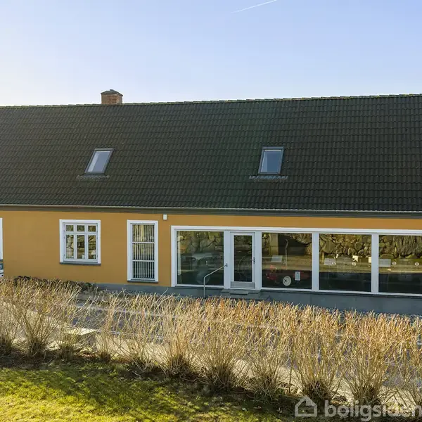 Et gult hus med mørkt tag har store vinduer foran. Det står i et åbent område med en lav hæk foran og en klar blå himmel i baggrunden.
