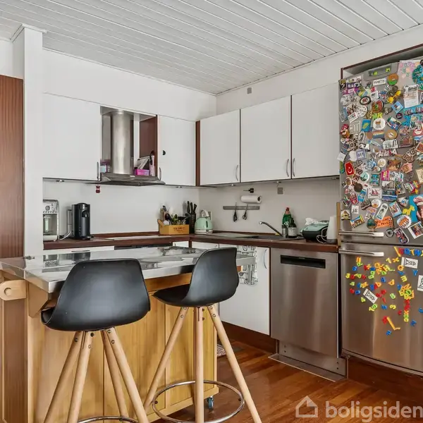 Et moderne køkken med en køkkenø udstyret med to høje, sorte stole. Køleskabet er dækket af farverige magneter. Rummet har hvide skabe og trægulv.