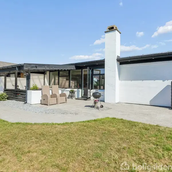 Et moderne, sort-hvidt hus med skorsten i en gårdhave med to liggestole og en grill. Omkring huset er der en velplejet græsplæne og en flisebelagt terrasse under en blå himmel.