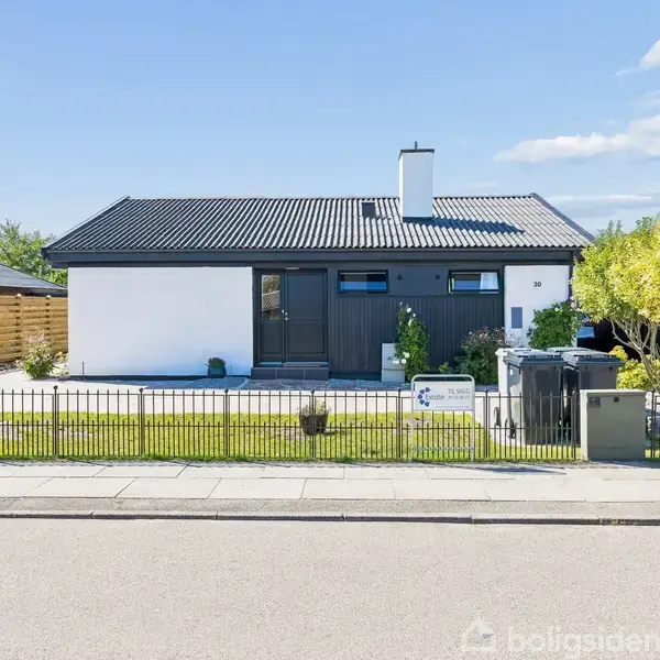 En moderne étplanshus med gråt tag og hvid facade står stille bag et sort hegn. En velplejet have omkranser huset, mens en klar blå himmel lyser op i baggrunden.