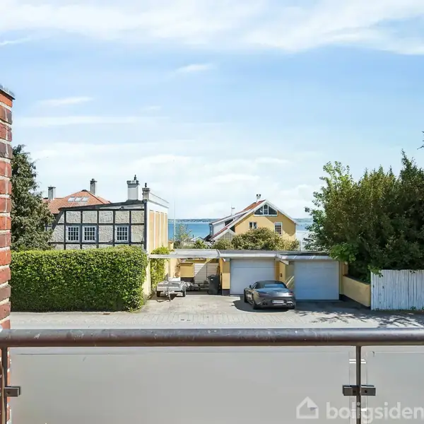 En balkon omgivet af en murstensvæg, med udsigt over en gårdsplads med parkerede biler, huse i baggrunden og havudsigt. Løvtræer omkranser gårdspladsen.