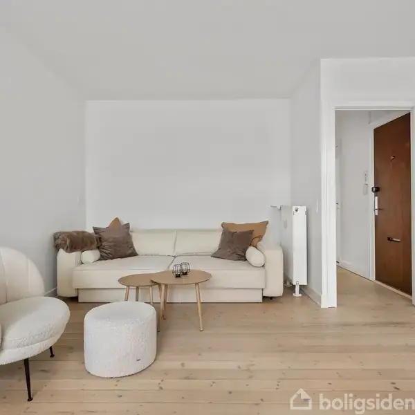 Sofa med puder står i en stue med trægulv, sammen med en rund stol og et lille bord. En åben dør leder til en gang.