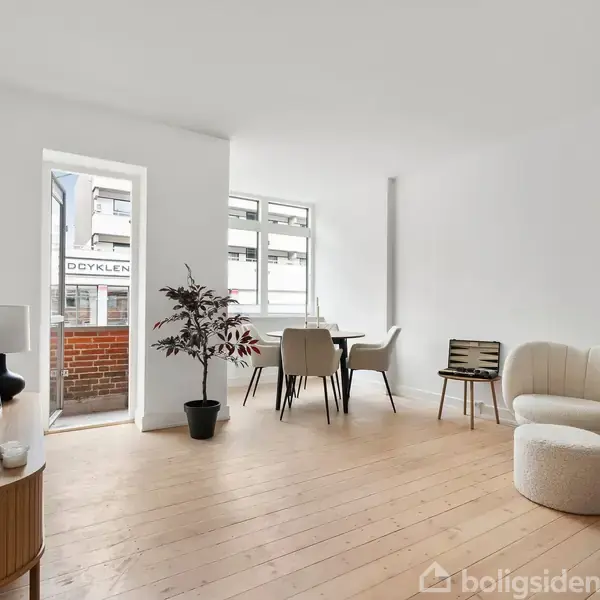 En lys stue indeholder et rundt bord med en stol og en sofa, placeret på et trægulv. En dør fører til en balkon, og et vindue giver naturligt lys.