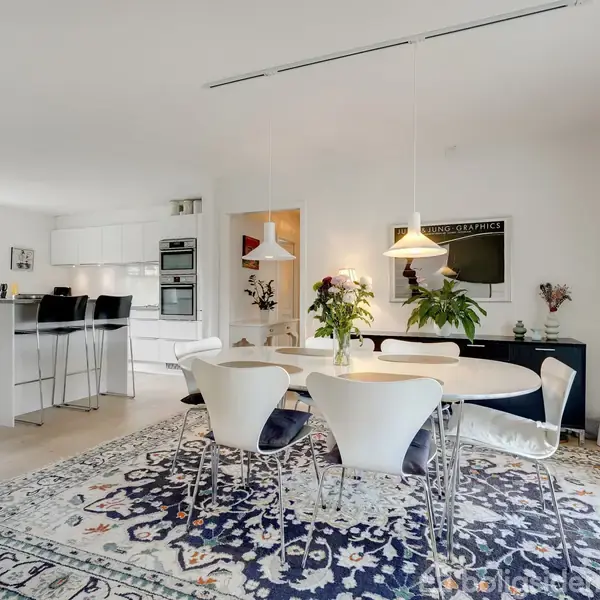 Spisebord med seks stole står på et mønstret tæppe i en lys stue. Over bordet hænger to lamper, og i baggrunden ses et moderne køkken med barstole.
