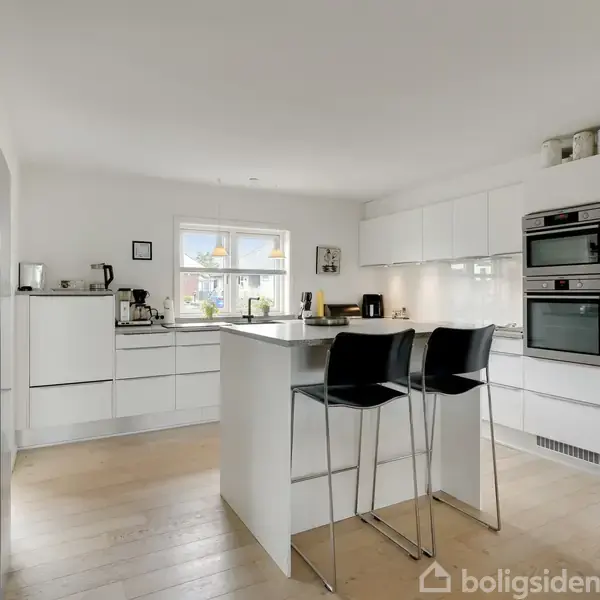 Et moderne, lyst køkken med hvide skabe og integrerede ovne. En køkkenø med to sorte barstole dominerer rummet. Vindue bringer naturligt lys ind, omgivet af træparketgulv.