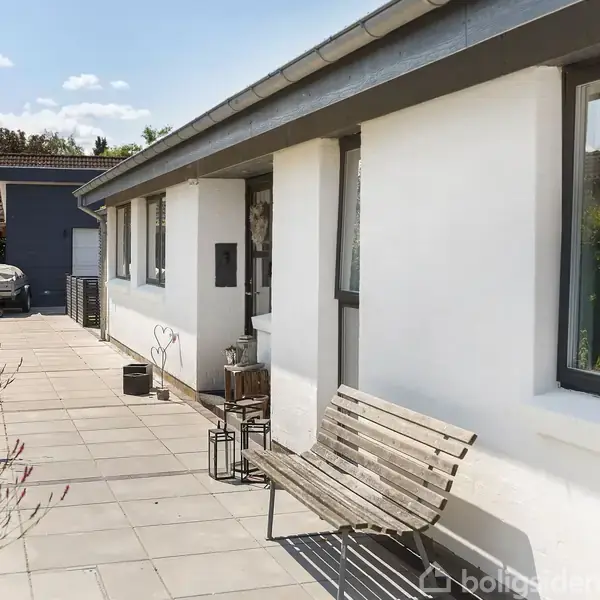 Hvidmalet hus med fladt tag og store vinduer står stille. En træbænk er placeret på den flisebelagte terrasse, omgivet af grønne buske. Klar blå himmel i baggrunden.