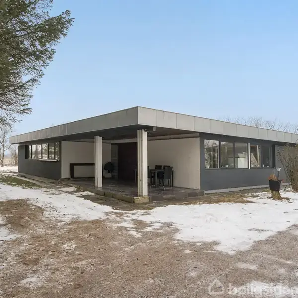 En enkeltstående, moderne bygning med fladt tag står i et vinterlandskab. Bygningen har store vinduer og en overdækket terrasse. Træer omgiver stedet, og jorden er delvist dækket af sne.