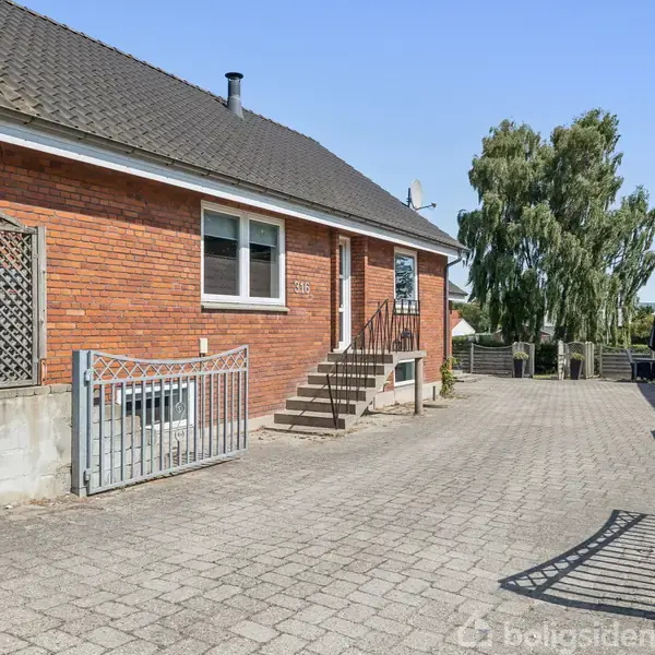 Rødt murstenshus med sort tegltag, adgang via trappe og metalgelænder. Brostensbelagt gårdsplads omgivet af hegn. Grønne træer i baggrunden samt blå himmel.