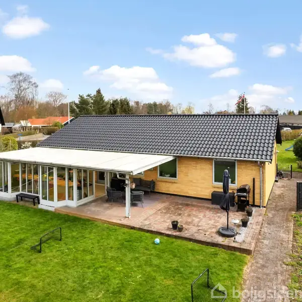 Et enkelt etplanshus står i en grøn have med en stor terrasse og en udestue. Omkringliggende træer og huse ses i baggrunden under en blå himmel med spredte skyer.