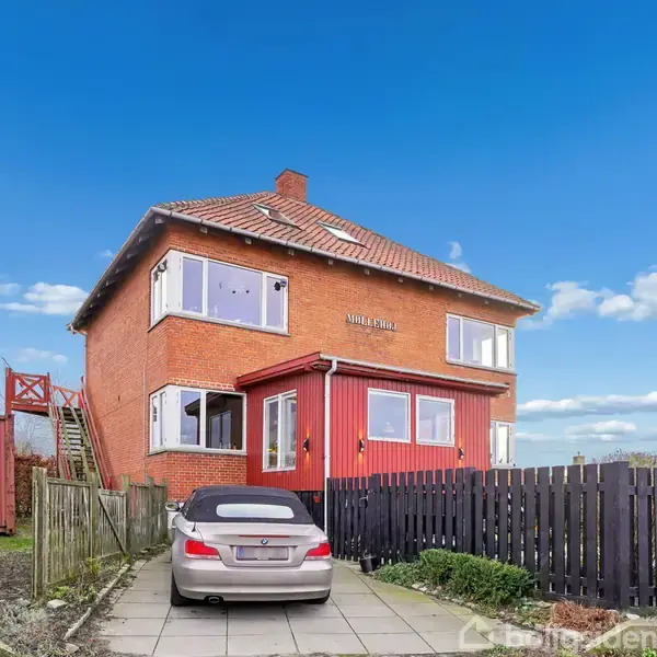 En hvid bygning med skråt tag er omgivet af frodig vegetation. Foran huset er en indhegnet havezone med et bord og stole, placeret midt i en græsfyldt have.