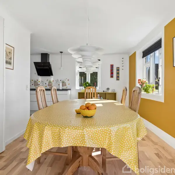 Et rundt spisebord er dækket med en gul prikket dug, skål med appelsiner ovenpå, omgivet af træstole i et lyst køkkenalrum med en gul accentvæg og store vinduer.