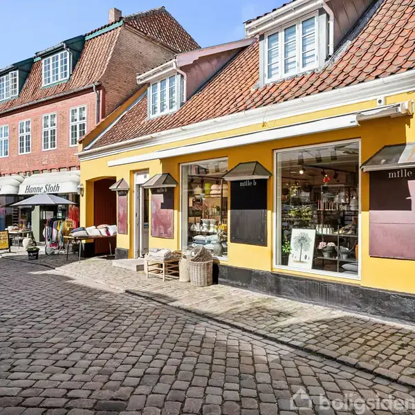 Butik med navnet "mille b" sælger varer fra store vinduer. Butikken ligger i et brostensbelagt gade miljø med flere butikker og røde murstenshuse omkring.