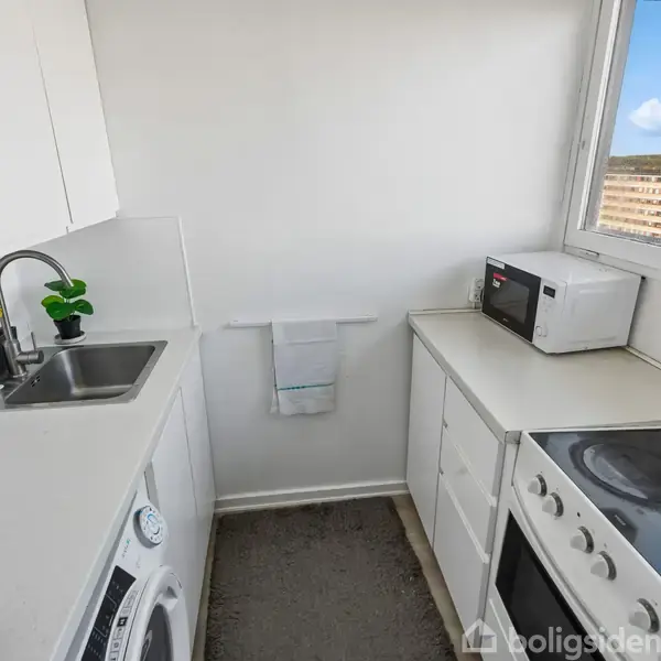Et lille køkken med en vaskemaskine, køkkenvask og komfur til højre. Mikrobølgeovn står på bordet. Hvidt interiør med vindue, der viser udsigt til andre bygninger.