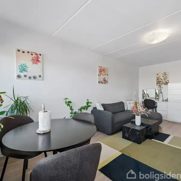 Et moderne, lyst stueinteriør med en grå sofa og sort sofabord. Rundt spisebord med stole og dekorative planter skaber en hyggelig atmosfære. To kunstværker hænger på væggen.