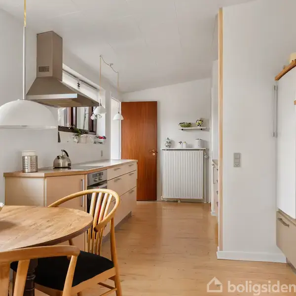 Spisebord med stole står i et lyst køkken. Køkkenet har træskabe, bordplade, hængende lamper og en dør til en anden del af huset. Vinduet giver udsyn til træer udenfor.