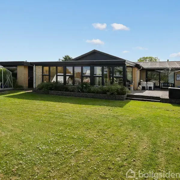 En moderne bungalow med store vinduer set forfra, omgivet af en velplejet græsplæne og et drivhus til venstre. Terrassen har udendørs møbler, og himlen er klar.