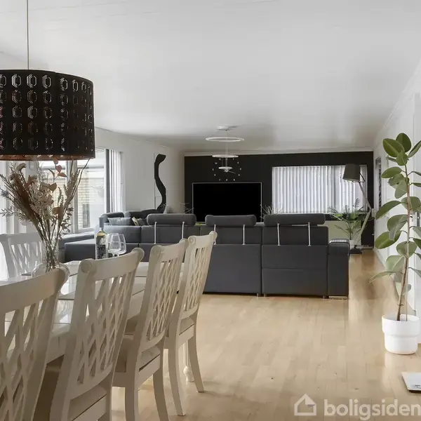 Spisebord med hvide stole står i en lys stue. Sofaer vender mod et fjernsyn. En stor, sort lampe hænger over bordet. En plante står ved siden af et vindue.