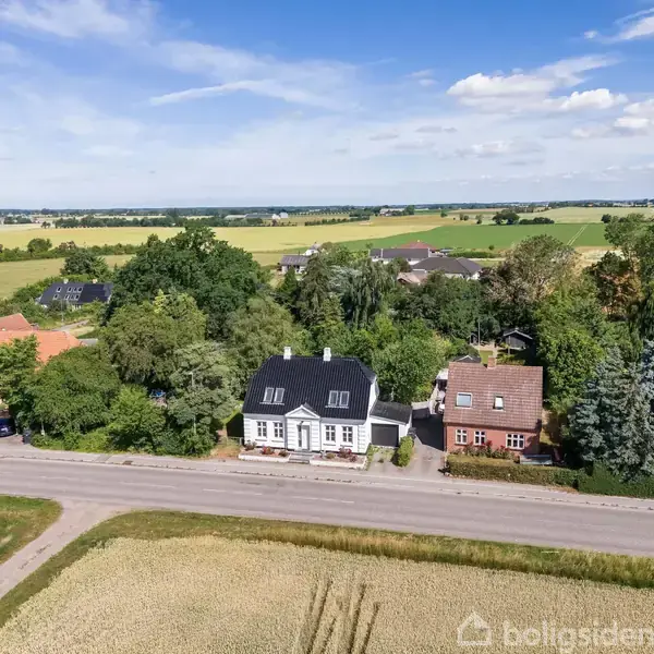 Flere huse står langs en vej omgivet af marker og træer. Himlen er blå med nogle skyer. Området er landligt med åbne marker i baggrunden.