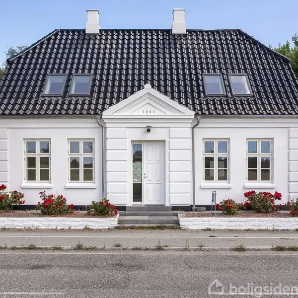 Et hvidt hus står stille langs en vej med et sort tegltag. Foran er der blomsterbede med røde blomster. Over døren står "1905". Træer omgiver huset.